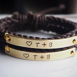 May include: Two brown braided cord bracelets with gold metal plates engraved with a heart and the text "T + B".
