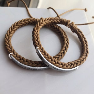 May include: Two brown braided bracelets with silver metal plates engraved with coordinates. The bracelets are tied together.
