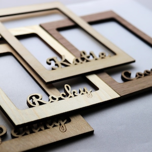 May include: A collection of rectangular wooden frames with laser-cut names attached to the bottom edges. The frames are made from light and dark wood tones, featuring the names Katie, Rocky, and Cooper in a cursive script font, layered over each other.