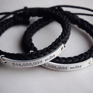May include: Two black braided bracelets with silver metal plates engraved with the text "584,000,000 miles" and "4,000,000 miles".