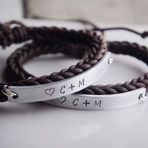May include: Two brown braided bracelets with silver metal plates engraved with the text "C + M" and a heart symbol.