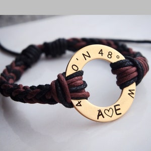 May include: A black and brown braided cord bracelet with a gold-toned metal ring charm. The charm has the inscription "O.N 48° A♥E M".