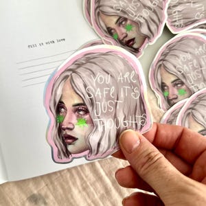 Könnte beinhalten: Ein gestanzter Aufkleber mit einer stilisierten Illustration einer Person mit welligem hellem Haar, rosafarbener Haut und grünen sternförmigen Markierungen unter den Augen. Der Text "YOU ARE SAFE IT'S JUST THOUGHTS" ist in Weiß darauf gedruckt.