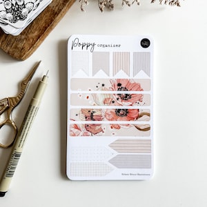 Könnte beinhalten: Ein weißes Poppy Organizer-Aufkleberblatt mit floralen Designs und bannerförmigen Aufklebern. Eine goldene Schere und ein Stift liegen neben dem Aufkleberblatt. Das Aufkleberblatt trägt den Text "Poppy Organizer" und "Melanie Bitters Illustrations".