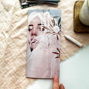 Könnte beinhalten: Ein rechteckiges Notizbuch mit einer gedruckten Illustration einer Person mit langem weißem Haar und ausdrucksstarken Augen, teilweise verdeckt durch detaillierte Strichzeichnungen von stacheligen Distelblüten in Braun- und Weißtönen.