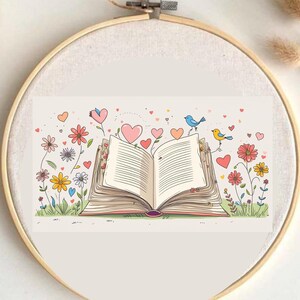 Puede incluir: Ilustración colorida de un libro abierto con corazones y flores. Dos pájaros se posan en las páginas. La obra de arte está dentro de un bastidor de bordado redondo de madera, creando una pieza decorativa.