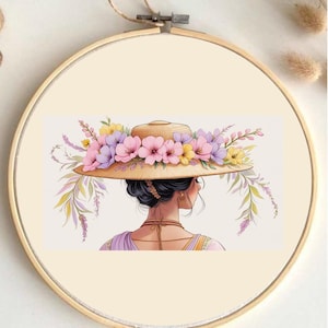 Puede incluir: Un bastidor de bordado con una ilustración en acuarela de una persona que lleva un sombrero de paja adornado con flores rosas, moradas y amarillas. La persona está de espaldas, con pelo oscuro y un collar.