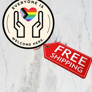 May include: A circular sticker with a cream background featuring a black line drawing of two hands cupping a multicoloured heart with the text EVERYONE IS WELCOME HERE. A red rectangular tag is attached to the side of the circular sticker.