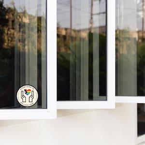 May include: A circular sticker featuring a black line drawing of two hands cupping a heart filled with the colours of the progress pride flag. The text "EVERYONE IS WELCOME HERE" is printed in black capital letters along the circular border.