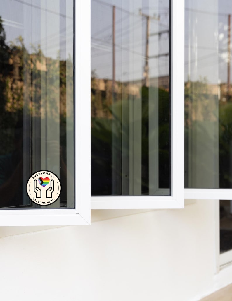 May include: A circular sticker featuring a black line drawing of two hands cupping a heart filled with the colours of the progress pride flag. The text "EVERYONE IS WELCOME HERE" is printed in black capital letters along the circular border.