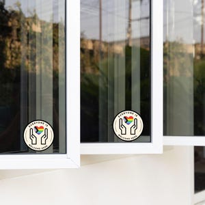 May include: Two circular stickers applied to window panes, each featuring a graphic of two hands cupping a rainbow-coloured heart. The text "EVERYONE IS WELCOME HERE" is printed in black capital letters along the circular border of each sticker.