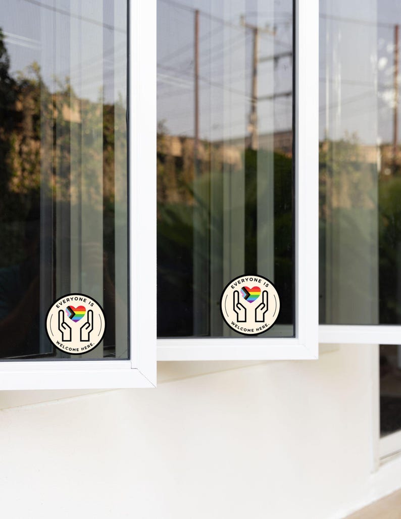 May include: Two circular stickers applied to window panes, each featuring a graphic of two hands cupping a rainbow-coloured heart. The text "EVERYONE IS WELCOME HERE" is printed in black capital letters along the circular border of each sticker.
