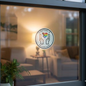 May include: A circular window decal featuring a black outline of two hands cupping a rainbow-coloured heart. The text "EVERYONE IS WELCOME HERE" is printed in black capital letters along the inner border of the circle on a white background.