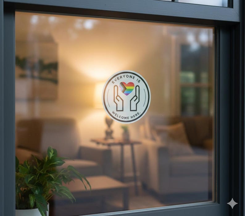 May include: A circular window decal featuring a black outline of two hands cupping a rainbow-coloured heart. The text "EVERYONE IS WELCOME HERE" is printed in black capital letters along the inner border of the circle on a white background.