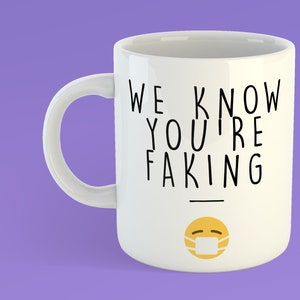 May include: White ceramic mug with black text that reads "We know you're faking" and a yellow emoji with a face mask.