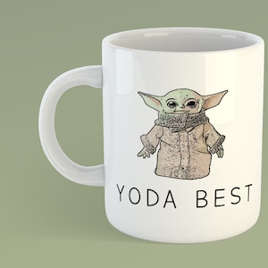 May include: White ceramic mug with a cartoon image of a green character wearing a brown coat and a scarf. The text "YODA BEST" is printed in black below the image.