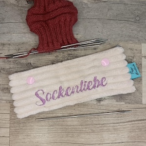 Könnte beinhalten: Ein weißes, geripptes Stoff-Nadelmäppchen mit dem lila gestickten Wort "Sockenliebe". Zwei rosa Knöpfe sichern das Etui. Eine teilweise gestrickte rote Socke und Stricknadeln sind ebenfalls zu sehen.
