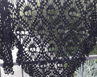 Può includere: Uno scialle nero fatto a mano all'uncinetto con un motivo floreale. Lo scialle è drappeggiato su una ringhiera di metallo, mostrando il suo design intricato e le delicate frange sui bordi. Il lavoro traforato crea un effetto pizzo.