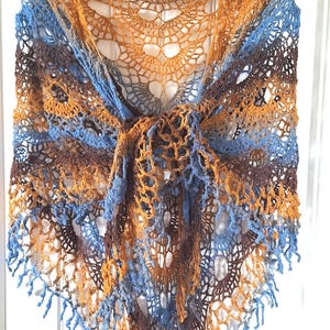May include: A triangular crochet shawl featuring an openwork lace pattern in a variegated color palette of brown, golden yellow, and light blue. The garment is finished with a delicate fringe along the bottom edges and is displayed on a hanger.