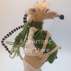 May include: A plush toy mouse wearing a green scarf and white pants, holding a green bag with a rope handle. The mouse has a black and white striped tail.