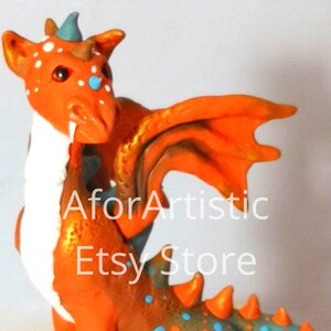 May include: A small orange and blue dragon figurine with white accents. The dragon has a long tail, wings, and a spiky back. It is sitting on a white surface.