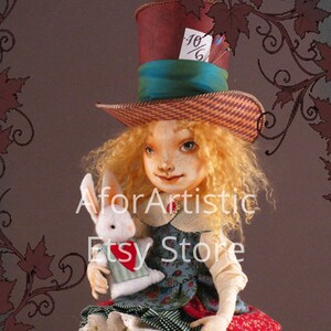 Op de afbeelding: Een handgemaakte pop gekleed als Alice uit Alice in Wonderland, met een roodbruine hoge hoed met een blauwe band en een wit label met de tekst "10/6". Ze houdt een wit konijn vast met een rood hart op zijn borst. De pop zit op een doos met een speelkaartenontwerp van de Koningin van harten.