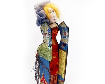 May include: A handmade doll with a blue and green patterned dress, a red skirt, and a blue head scarf. The doll is holding a blue and yellow patterned book.