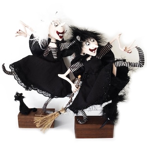 May include: Two black-dressed witches with white and black striped shirts, one with white hair and the other with black hair, are flying on broomsticks. A black cat is sitting on a wooden base.