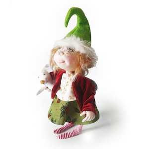 May include: A handmade Christmas elf doll with a green hat, red jacket, and green skirt with holly berry print. The doll is holding a white animal and has striped red and white socks. The doll has freckles and a smiling expression.