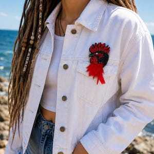 May include: A white denim jacket featuring a beaded rooster brooch on the chest pocket. The brooch is crafted with red and black beads, a white eye, and long red feathers extending downward from the base of the rooster figure.