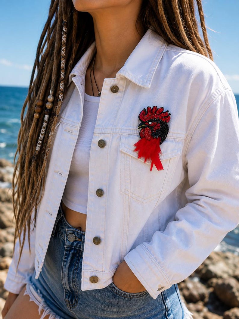 May include: A white denim jacket featuring a beaded rooster brooch on the chest pocket. The brooch is crafted with red and black beads, a white eye, and long red feathers extending downward from the base of the rooster figure.