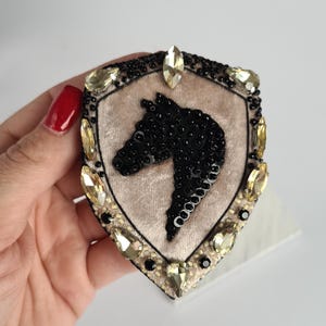 Op de afbeelding: Een schildvormige broche met een paardenkopontwerp. De broche heeft een beige fluwelen midden met een zwarte, met kralen bezette paardenkop. De rand is versierd met zwarte kralen en geel getinte kristallen. Een decoratief accessoire.