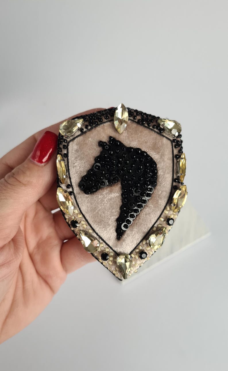 Op de afbeelding: Een schildvormige broche met een paardenkopontwerp. De broche heeft een beige fluwelen midden met een zwarte, met kralen bezette paardenkop. De rand is versierd met zwarte kralen en geel getinte kristallen. Een decoratief accessoire.