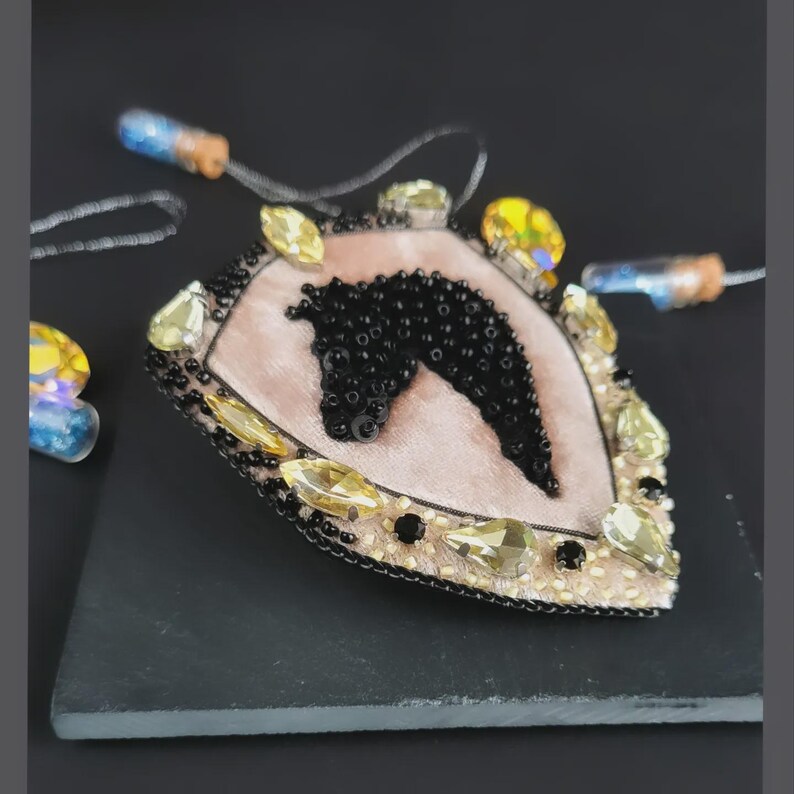 Op de afbeelding: Handgemaakte broche met een paardenhoofd ontwerp. De broche heeft een roze fluwelen midden met een zwart, met kralen bezet paardenhoofd. Het is omlijst met goudkleurige kralen en gele kristallen. De broche ligt op een zwart oppervlak.