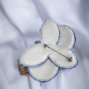May include: A butterfly-shaped brooch featuring white textured faux leather petals with a blue beaded edge. The back of the brooch displays a silver-toned metal pin mechanism attached to the center, allowing for secure fastening to various garments.