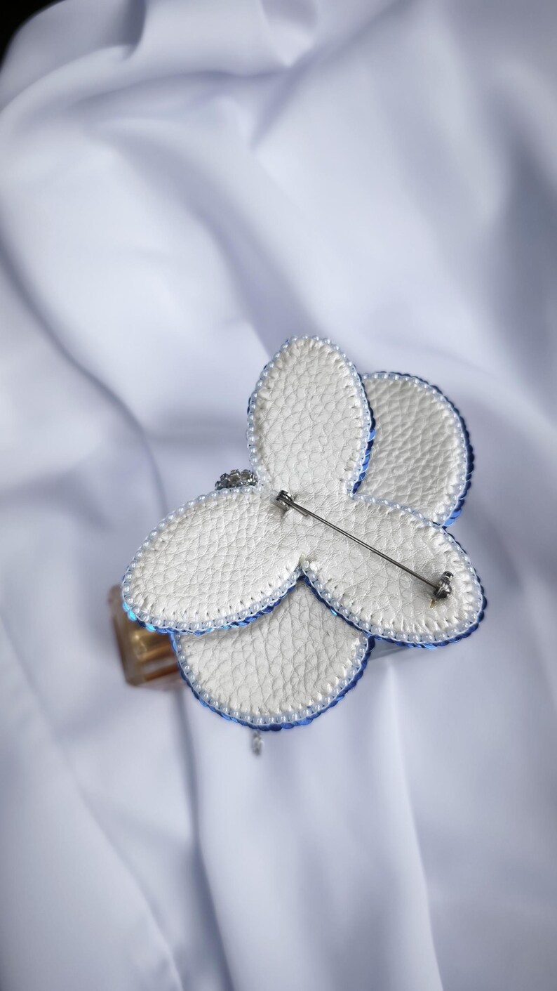 May include: A butterfly-shaped brooch featuring white textured faux leather petals with a blue beaded edge. The back of the brooch displays a silver-toned metal pin mechanism attached to the center, allowing for secure fastening to various garments.