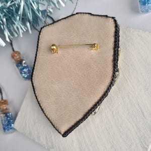 Op de afbeelding: Een schildvormige broche met een tan leren achterkant en een zwarte gevlochten rand. De broche heeft een gouden speldsluiting en is versierd met zilveren en heldere kristallen. Kleine glazen flesjes gevuld met blauwe glitter op de achtergrond.