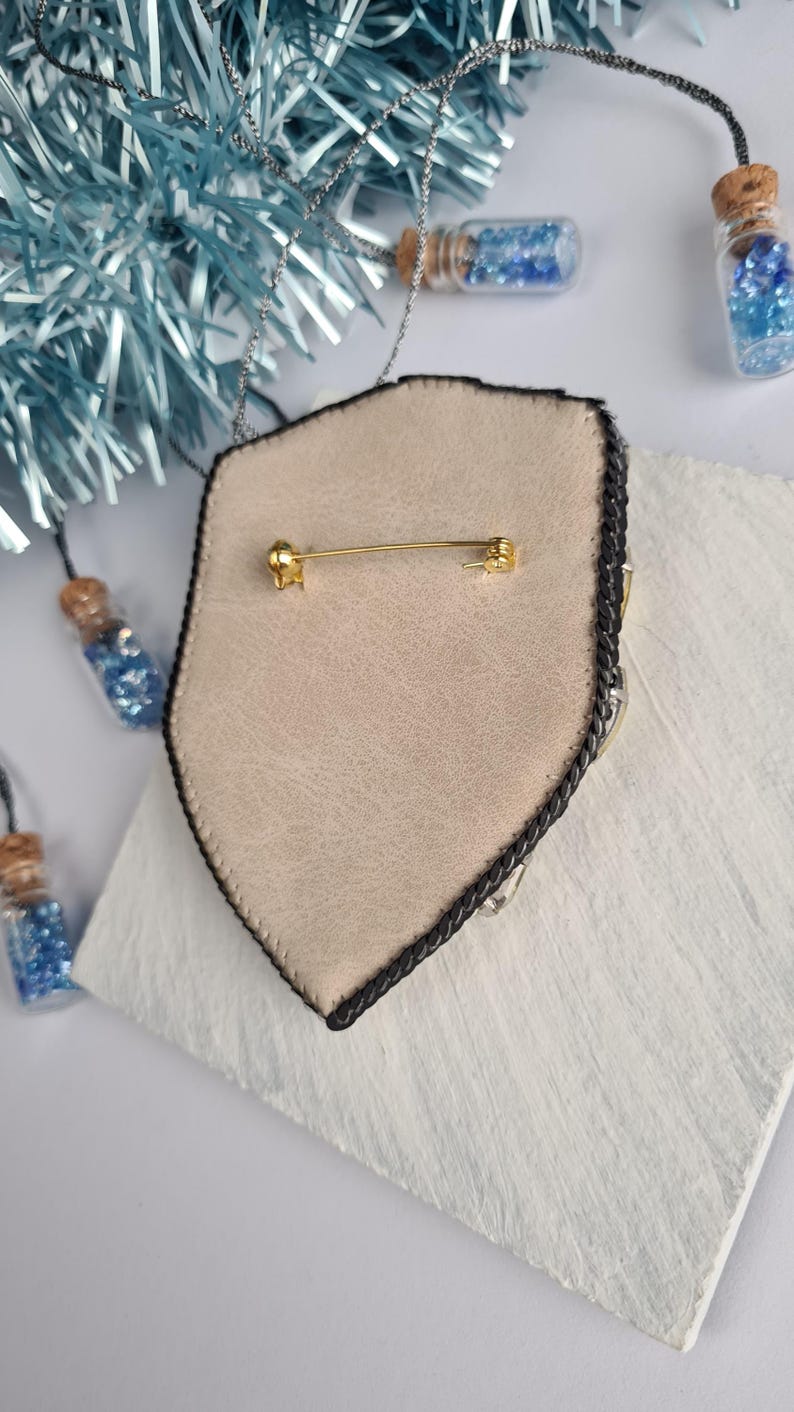 Op de afbeelding: Een schildvormige broche met een tan leren achterkant en een zwarte gevlochten rand. De broche heeft een gouden speldsluiting en is versierd met zilveren en heldere kristallen. Kleine glazen flesjes gevuld met blauwe glitter op de achtergrond.