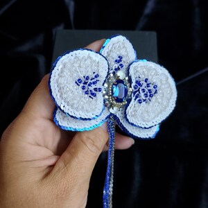May include: A handcrafted orchid brooch featuring white and blue sequins, small white beads, and a central oval blue gemstone. The flower design includes a dangling beaded fringe in blue and silver tones, creating a detailed floral accessory.