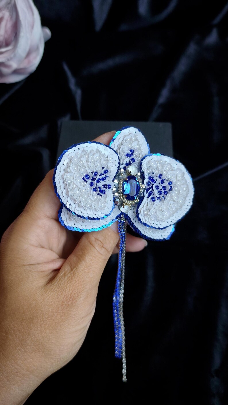 May include: A handcrafted orchid brooch featuring white and blue sequins, small white beads, and a central oval blue gemstone. The flower design includes a dangling beaded fringe in blue and silver tones, creating a detailed floral accessory.