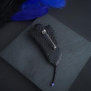 May include: A black leather flower brooch with a blue bead at the end of the pin. The brooch is on a black slate surface.