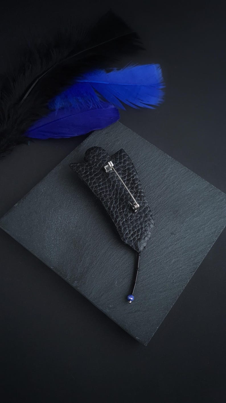 May include: A black leather flower brooch with a blue bead at the end of the pin. The brooch is on a black slate surface.