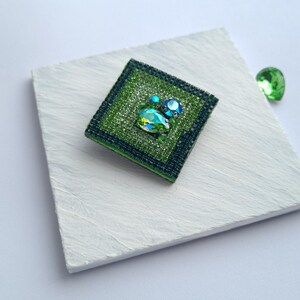 May include: A square, green beaded brooch with a central teardrop-shaped crystal and two smaller blue crystals. The brooch is set on a white surface, with a loose green crystal nearby. The brooch has a dark green border.