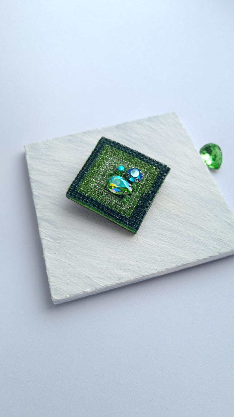 May include: A square, green beaded brooch with a central teardrop-shaped crystal and two smaller blue crystals. The brooch is set on a white surface, with a loose green crystal nearby. The brooch has a dark green border.