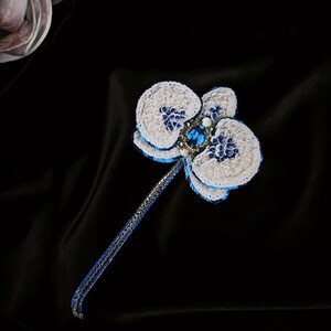 May include: A handcrafted orchid brooch featuring white petals with blue sequin accents and a central oval blue gemstone. The flower is attached to a long, slender stem made of blue and silver metallic thread, creating a detailed floral accessory.