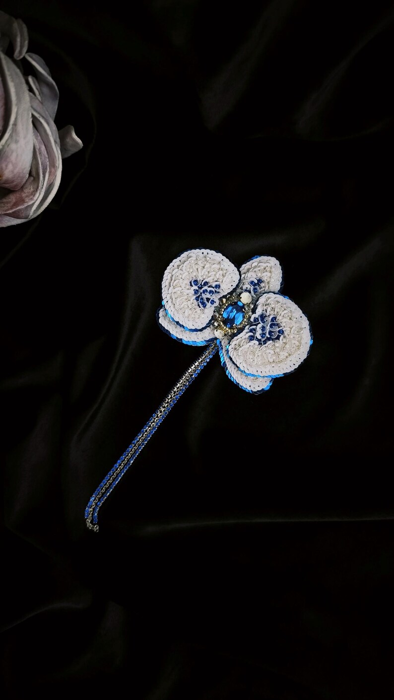 May include: A handcrafted orchid brooch featuring white petals with blue sequin accents and a central oval blue gemstone. The flower is attached to a long, slender stem made of blue and silver metallic thread, creating a detailed floral accessory.