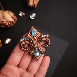 Op de afbeelding: Een gedetailleerde, handgemaakte fleur-de-lis broche. De broche heeft een koperkleurige basis, versierd met kleine, ronde kralen en sprankelende blauwe en heldere kristallen. Het ontwerp is ingewikkeld en elegant, geschikt voor formele kleding.
