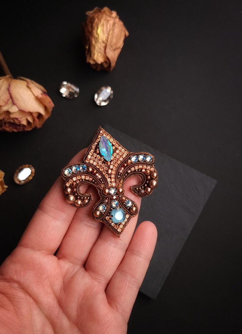 Op de afbeelding: Een gedetailleerde, handgemaakte fleur-de-lis broche. De broche heeft een koperkleurige basis, versierd met kleine, ronde kralen en sprankelende blauwe en heldere kristallen. Het ontwerp is ingewikkeld en elegant, geschikt voor formele kleding.