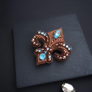 May include: A detailed, handcrafted brooch in the shape of a fleur-de-lis. The brooch is embellished with copper-colored beads and light blue gemstones. The design is set against a dark background, with a loose gemstone nearby.
