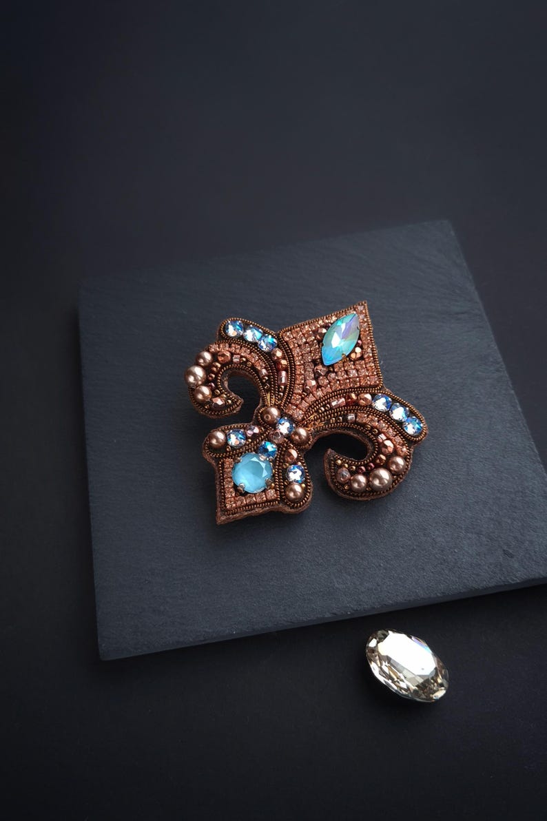 Op de afbeelding: Een gedetailleerde, handgemaakte broche in de vorm van een fleur-de-lis. De broche is versierd met koperkleurige kralen en lichtblauwe edelstenen. Het ontwerp is geplaatst tegen een donkere achtergrond, met een losse edelsteen in de buurt.